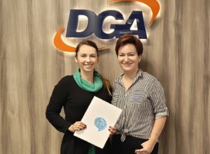 Biuro z&nbsp;neonem DGA. Dwie kobiety trzymają teczkę z&nbsp;podpisanymi dokumentami. Uśmiechają się.